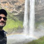 Selfie de um homem em frente ao pé de uma cascata, com arco-iris