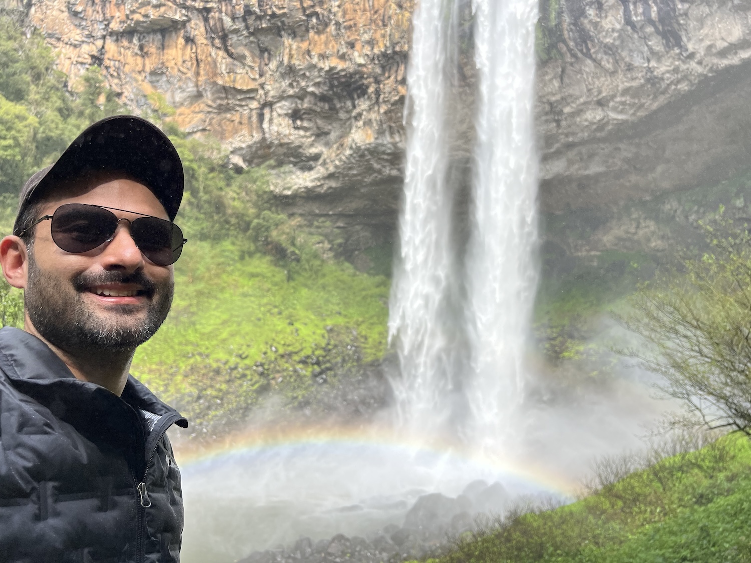 Selfie de um homem em frente ao pé de uma cascata, com arco-iris
