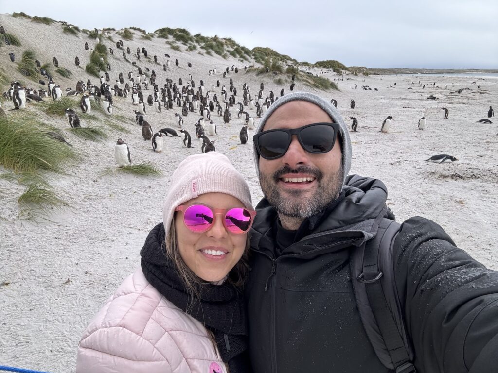 Casal fazendo selfie com colônia de pinguins ao fundo. 