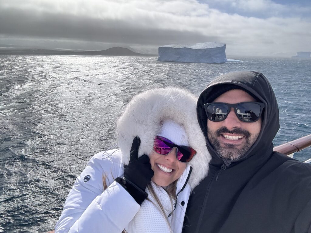 casal fazendo selfie em frente a um iceberg, com dia ensolarado. 