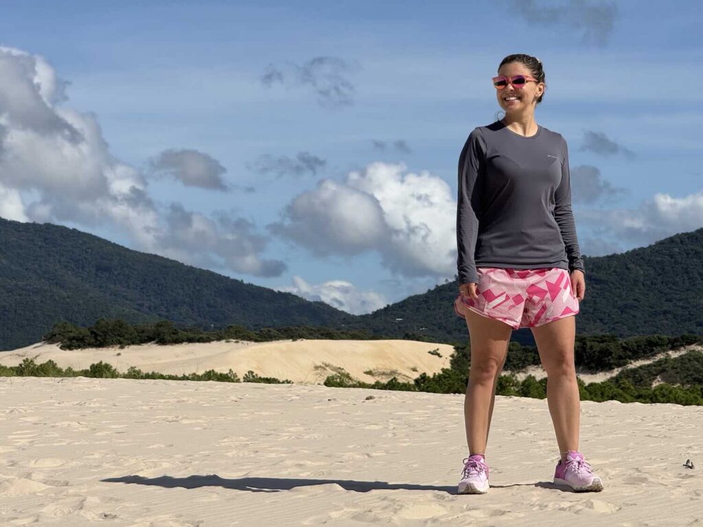 mulher com roupa esportiva, céu azul com nuvens, na areia com morros de vegetação ao fundo