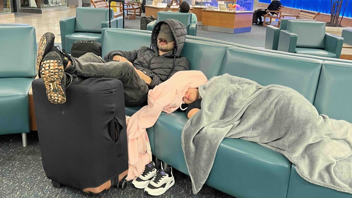 Homem e mulher dormindo no banco de um aeroporto com malas e cobertas