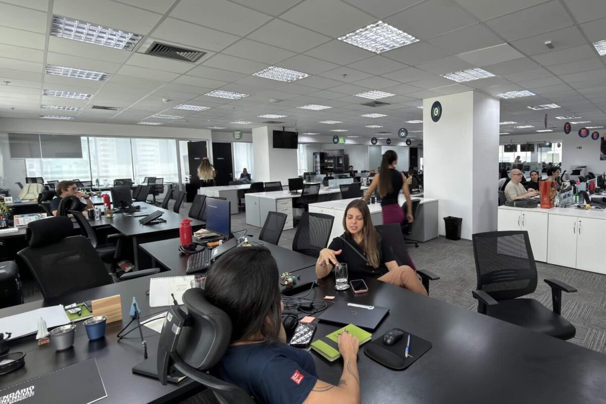 mulheres sentadas conversando em um escritório com várias pessoas nas mesas de trabalho.